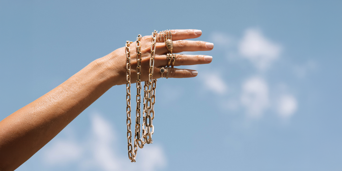 Hand wearing multiple gold bracelets and rings against a blue sky background